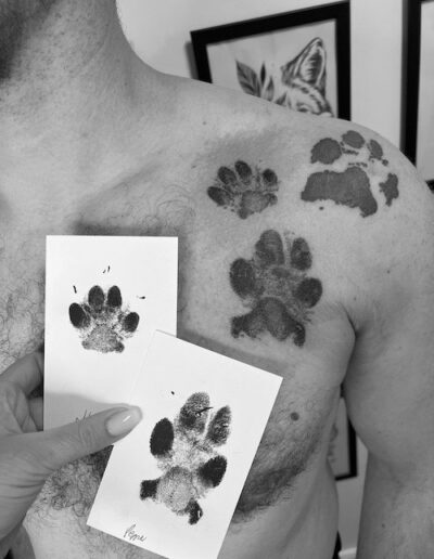Schwarz-weiß Fotografie einer Schulter mit mehreren tätowierten Pfotenabdrücken, daneben werden Papierkarten mit originalen Pfotenabdrücken gehalten.